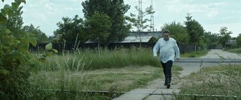 Movie still from “Graduation” (2016), directed by Cristian Mungiu – A man walking down a sidewalk near a field; Wide shot, Low angle