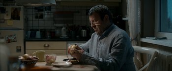 Movie still from “Graduation” (2016), directed by Cristian Mungiu – A man sitting at a table eating a sandwich; Close Up shot, Over the shoulder angle