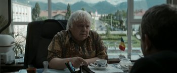 Movie still from “Graduation” (2016), directed by Cristian Mungiu – An older man sitting at a table with a cup of coffee; Medium shot, Over the shoulder angle