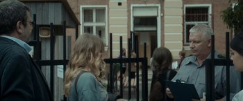 Movie still from “Graduation” (2016), directed by Cristian Mungiu – A woman standing in front of a building with a fence; Medium shot, Over the shoulder angle