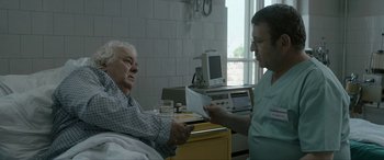 Movie still from “Graduation” (2016), directed by Cristian Mungiu – An older man in a hospital bed holding a paper; Medium shot, Over the shoulder angle