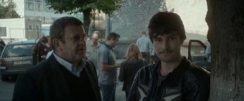 Movie still from “Graduation” (2016), directed by Cristian Mungiu – Two men standing next to each other on a sidewalk; Close Up shot, Over the shoulder angle