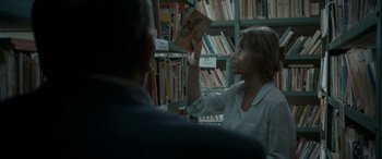Movie still from “Graduation” (2016), directed by Cristian Mungiu – A woman in white shirt looking at a book in a library; Medium shot, Over the shoulder angle