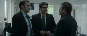 Movie still from “Graduation” (2016), directed by Cristian Mungiu – A group of men standing next to each other in a room; Medium shot, Over the shoulder angle