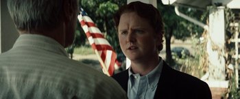 Movie still from “Gran Torino” (2008), directed by Clint Eastwood – A man in a suit and tie standing next to an american flag; Close Up shot, Over the shoulder angle