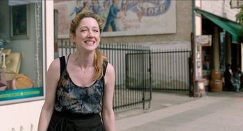 Movie still from “Grandma” (2015), directed by Paul Weitz – A woman standing on the sidewalk smiling for the camera; Medium shot, Over the shoulder angle