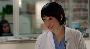 Movie still from “Grandma” (2015), directed by Paul Weitz – A woman wearing a white lab coat smiling for the camera; Close Up shot, Over the shoulder angle