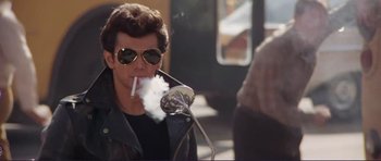 Movie still from “Grease 2” (1982), directed by Patricia Birch – A man in sunglasses smoking a cigarette on the street; Medium shot, Low angle