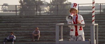 Movie still from “Grease 2” (1982), directed by Patricia Birch – A man in a cheerleader uniform standing on a bleacher with a megaphone; Wide shot, Low angle