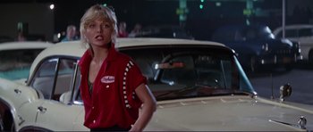 Movie still from “Grease 2” (1982), directed by Patricia Birch – A woman standing next to a car in a parking lot at night; Medium shot, Low angle