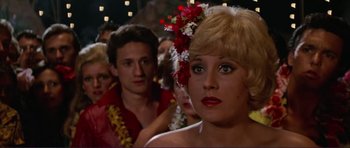 Movie still from “Grease 2” (1982), directed by Patricia Birch – A woman with a flower in her blonde hair and a man with a flower in his hair in the background; Close Up shot, Over the shoulder angle