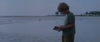 Movie still from “Great Expectations” (1998), directed by Alfonso Cuarón – A young boy writing on a piece of paper by a body of water; Medium shot, Low angle
