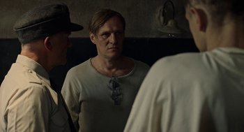 Movie still from “Great Freedom” (2021), directed by Sebastian Meise – A man in a white t - shirt with a tie around his neck looks into a mirror; Medium shot, Over the shoulder angle