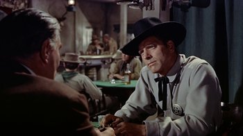 Movie still from “Gunfight at the O.K. Corral” (1957), directed by John Sturges – A man sitting at a table in a room with other people; Medium shot, Over the shoulder angle