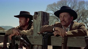 Movie still from “Gunfight at the O.K. Corral” (1957), directed by John Sturges – Two men in cowboy hats leaning on a wooden fence; Medium shot, Low angle