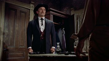 Movie still from “Gunfight at the O.K. Corral” (1957), directed by John Sturges – A man in a suit and hat standing in front of a table; Medium shot, Low angle