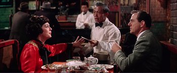 Movie still from “Gypsy” (1962), directed by Mervyn LeRoy – An older man is holding a piece of paper in front of a group of people at a table; Medium shot, Over the shoulder angle