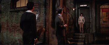 Movie still from “Gypsy” (1962), directed by Mervyn LeRoy – A man in a suit and a hat standing in front of a brick wall; Wide shot, Over the shoulder angle