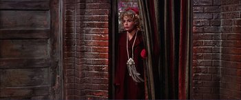 Movie still from “Gypsy” (1962), directed by Mervyn LeRoy – A woman in a red coat standing in front of a brick wall; Medium shot, Low angle