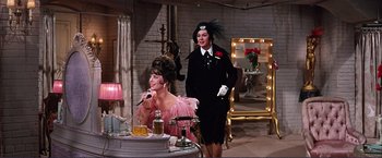 Movie still from “Gypsy” (1962), directed by Mervyn LeRoy – A woman in a pink dress and a man in a black suit stand next to a table; Medium shot, High angle