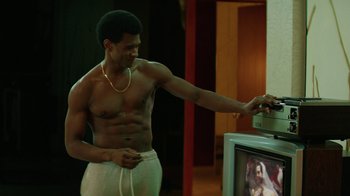 Movie still from “Hands of Stone” (2016), directed by Jonathan Jakubowicz – A shirtless man standing in front of a television; Medium shot, Over the shoulder angle