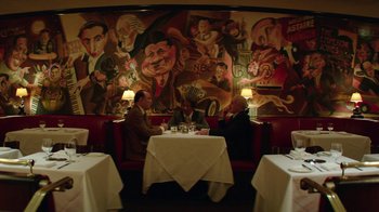 Movie still from “Hands of Stone” (2016), directed by Jonathan Jakubowicz – Two men sitting at a table in front of a painting; Wide shot, High angle
