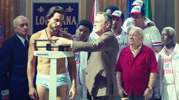 Movie still from “Hands of Stone” (2016), directed by Jonathan Jakubowicz – A group of people standing around a man in a suit; Medium shot, Over the shoulder angle