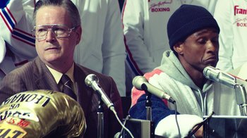 Movie still from “Hands of Stone” (2016), directed by Jonathan Jakubowicz – Two men are sitting at a podium with microphones; Close Up shot, Over the shoulder angle