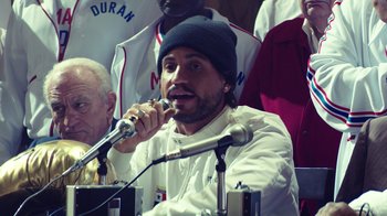Movie still from “Hands of Stone” (2016), directed by Jonathan Jakubowicz – A man in a white shirt and a black hat is speaking; Close Up shot, Over the shoulder angle