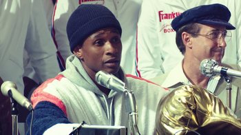 Movie still from “Hands of Stone” (2016), directed by Jonathan Jakubowicz – A man with a microphone in front of a group of people; Close Up shot, Over the shoulder angle