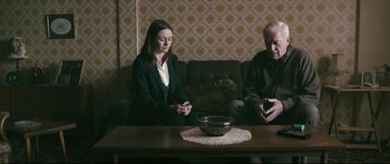 Movie still from “Harry Brown” (2009), directed by Daniel Barber – A woman sitting on a couch next to an older man; Medium shot, Over the shoulder angle