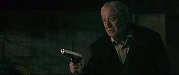 Movie still from “Harry Brown” (2009), directed by Daniel Barber – An older man holding a gun in a dark room; Medium shot, Over the shoulder angle