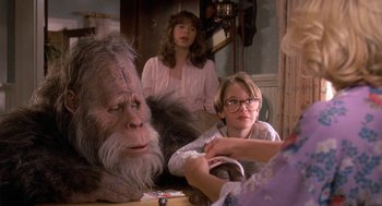 Movie still from “Harry and the Hendersons” (1987), directed by William Dear – A group of people sitting around a table with a stuffed animal; Medium shot, Over the shoulder angle