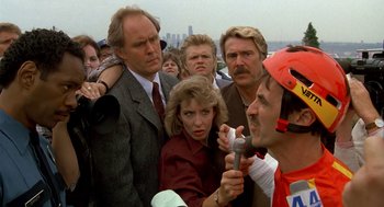 Movie still from “Harry and the Hendersons” (1987), directed by William Dear – A group of people gathered around a microphone; Close Up shot, Over the shoulder angle