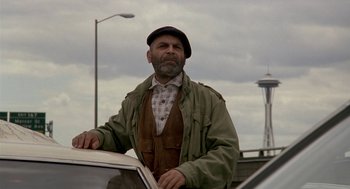 Movie still from “Harry and the Hendersons” (1987), directed by William Dear – An older man standing next to a car in a parking lot; Medium shot, Low angle