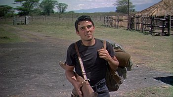 Movie still from “Hatari!” (1962), directed by Howard Hawks – A man holding a rifle and a backpack on the side of a dirt road; Medium shot, Low angle