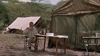 Movie still from “Hatari!” (1962), directed by Howard Hawks – A man sitting at a table in front of a tent; Wide shot, Over the shoulder angle