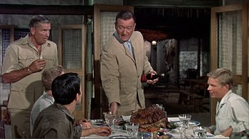 Movie still from “Hatari!” (1962), directed by Howard Hawks – A man in a tan jacket standing at a table with food; Medium shot, Over the shoulder angle