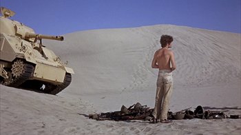 Movie still from “Head” (1968), directed by Bob Rafelson – A man standing on top of a sandy beach next to a tank; Wide shot, Low angle