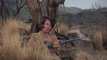 Movie still from “Head” (1968), directed by Bob Rafelson – A man sitting in the grass holding a rifle; Medium shot, Low angle