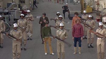 Movie still from “Head” (1968), directed by Bob Rafelson – A group of people standing on a street holding baseball bats; Wide shot, High angle