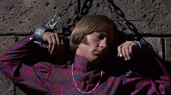 Movie still from “Head” (1968), directed by Bob Rafelson – A man in a colorful shirt is chained to a wall; Medium shot, High angle