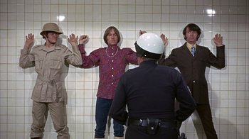 Movie still from “Head” (1968), directed by Bob Rafelson – A group of people standing in a room with a police officer standing in front of them; Medium shot, Low angle
