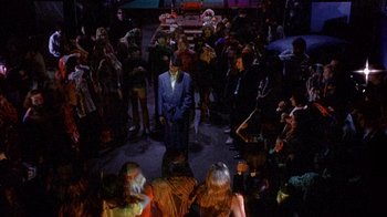 Movie still from “Head” (1968), directed by Bob Rafelson – A crowd of people sitting in a room with a man in a blue suit; Wide shot, High angle