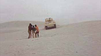 Movie still from “Head” (1968), directed by Bob Rafelson – Three people standing in the sand looking at a tank; Extreme Wide shot, High angle