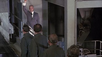 Movie still from “Head” (1968), directed by Bob Rafelson – A man in a suit and tie standing in front of a group of people; Wide shot, Over the shoulder angle