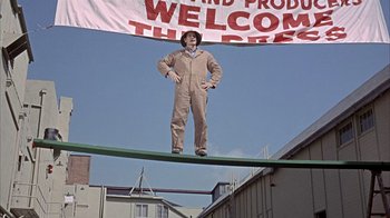 Movie still from “Head” (1968), directed by Bob Rafelson – A man standing on top of a pole under a banner; Wide shot, Low angle