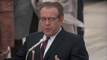 Movie still from “Head” (1968), directed by Bob Rafelson – An older man in a suit and tie speaking into a microphone; Close Up shot, Over the shoulder angle