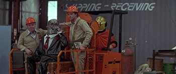 Movie still from “Heartbeeps” (1981), directed by Allan Arkush – A man standing next to another man on a forklift; Medium shot, Low angle