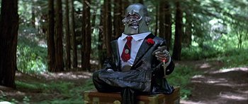 Movie still from “Heartbeeps” (1981), directed by Allan Arkush – A statue of a man in a suit sitting on a bench in the woods; Medium shot, Low angle